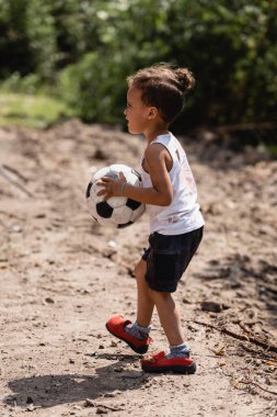 Gecekondu mahallesinde futbol topu tutan yoksul Afro-Amerikan çocuğun yan görüntüsü. 