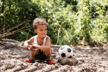 Pis suratlı, gülümseyen Afrikalı Amerikalı çocuk kirli bir yolda futbolun yanında tahta dal tutuyor. 