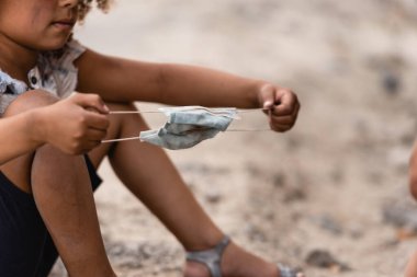 Cropped view of homeless african american child holding dirty medical mask on urban street  clipart