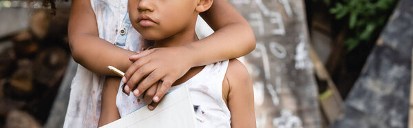 panoramic crop of african american kid holding pencil and hugging poor brother in torn clothes 