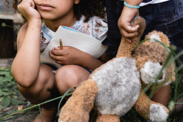 cropped view of poor african american kid holding notebook near child standing with dirty soft toy