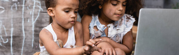 panoramic crop of poor african american children looking at laptop outside 