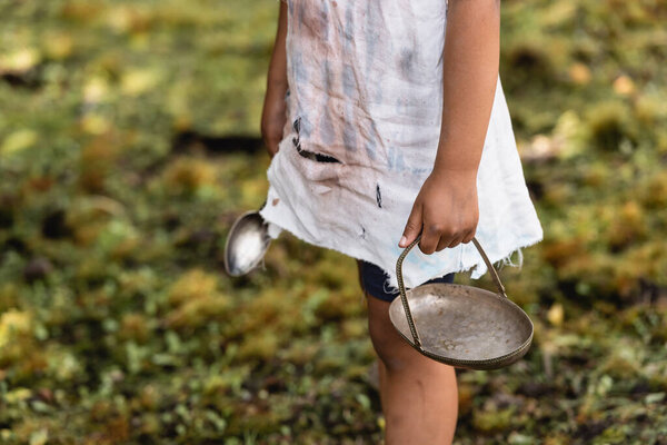 Cropped view of poor african american child holding metal plate and spoon on urban street 