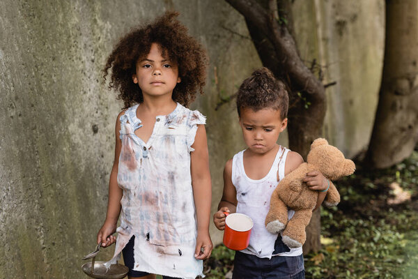 Poor arican American kid holding metal spoon and plate near sad brother with dirty teddy bear and cup in slum 