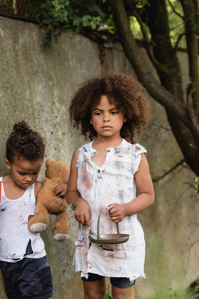 Dirty african american children with metal plate and dirty teddy bear standing in slum 