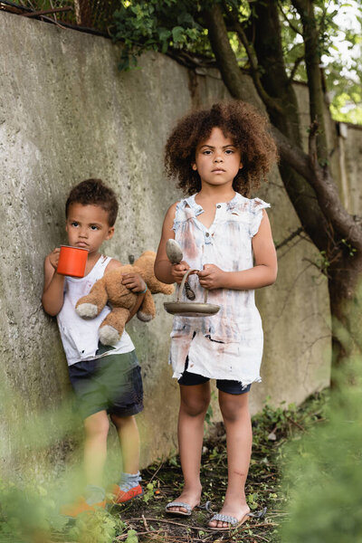 Selective focus of poor african american child holding metal plate and spoon near brother with cup and dirty teddy bear near concrete wall on urban street 