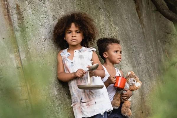 貧しいアフリカ系アメリカ人の子供の選択的な焦点金属スプーンとプレート近くの弟とともにカップと汚いテディベアでスラム — ストック写真