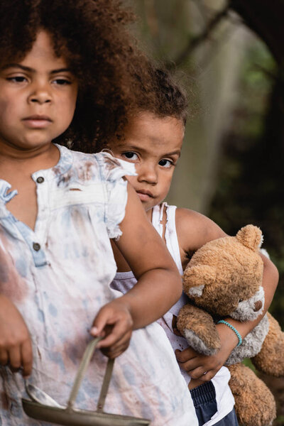 Selective focus of poor african american boy holding messy teddy bear near sister with metal plate on urban street 