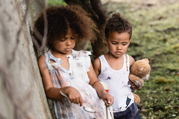 Selective focus of helpless african american kids with spoon, cup and teddy bear standing near concrete wall on urban street 
