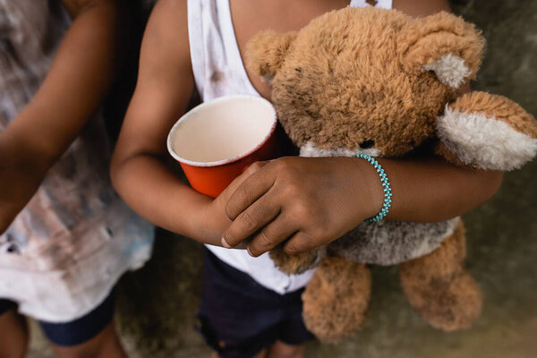 Cropped view of dirty african american children with teddy bear and cup begging alms on urban street 