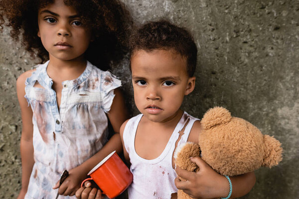 Poor african american children in dirty clothes with metal cup and teddy bear looking at camera near concrete wall outdoors 