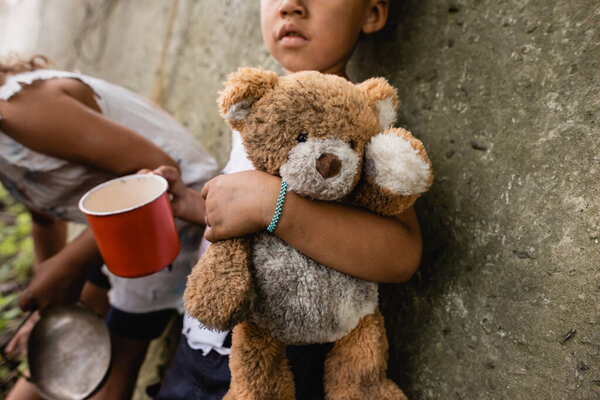 Cropped view of homeless african american children with dirty teddy bear begging alms in slum 