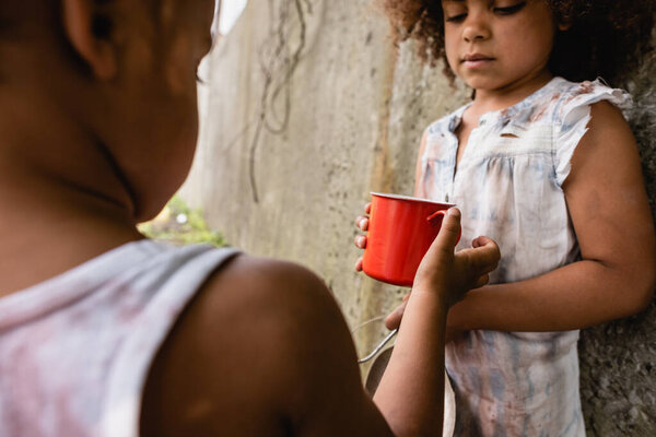 Selective focus of african american kid giving metal cup to sister in dirty clothes on urban street 