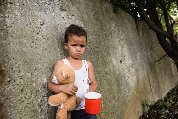Displeased african american boy holding dirty teddy bear and metal cup near concrete wall on urban street 