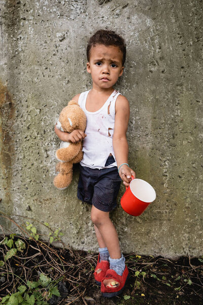 Dissatisfied african american child in dirty clothes begging alms near concrete wall in slum 