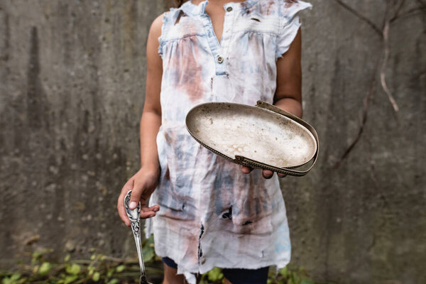 Cropped view of poor african american child holding dirty metal plate while begging alms on urban street 