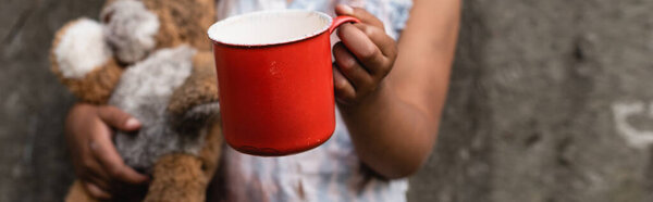 Panoramic orientation of poor african american child holding metal cup for alms on urban street 