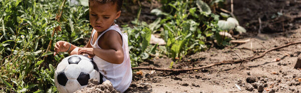 Панорамный урожай бедного африканского мальчика, сидящего рядом с футболом на грязной дороге на городской улице 