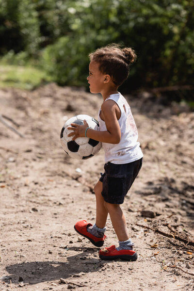 Side view of destitute african american boy holding soccer ball on dirty road in slum 
