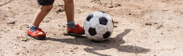 Panoramic crop of african american kid playing football on dirty road in slum 
