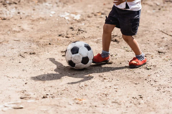 Cropped view of poor african american boy playing with soccer ball on dirty road on urban street 