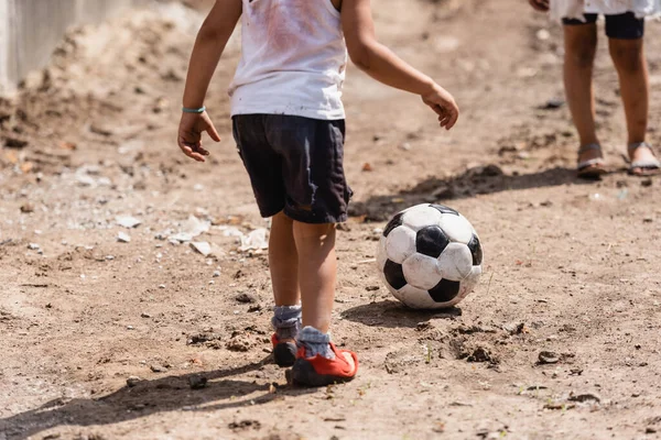 Cropped view of poor african american boy playing football near sister on dirty road in slum 