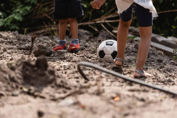 Cropped view of poor african american children playing football on dirty road in slum 