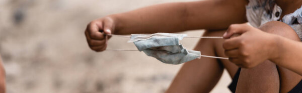 Panoramic orientation of poor african american kid holding messy medical mask on urban street 