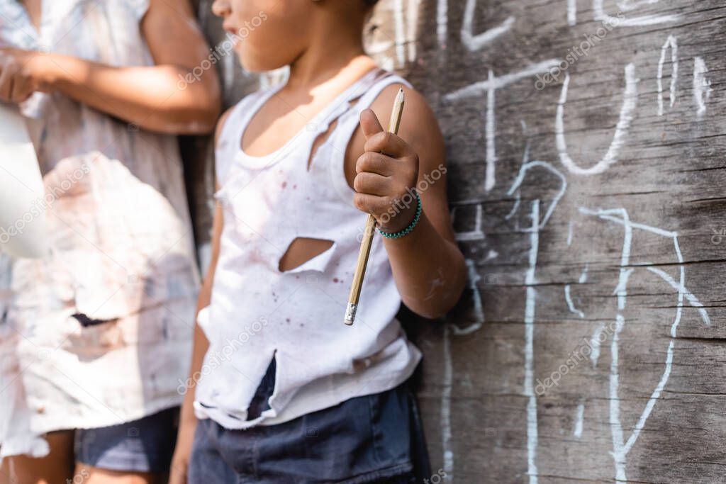 cropped view of poor african american kid in torn clothes standing near chalkboard and sister while holding pencil 