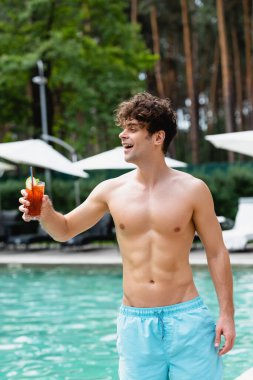 cheerful and shirtless man holding glass with cocktail near swimming pool clipart