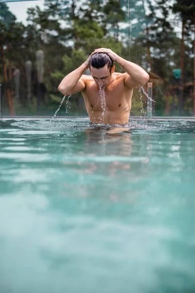 selective focus of wet and muscular man touching hair in swimming pool