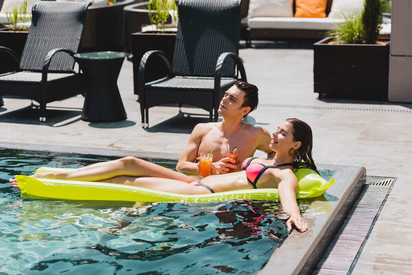 beautiful woman lying on inflatable mattress near handsome man holding cocktail in pool 