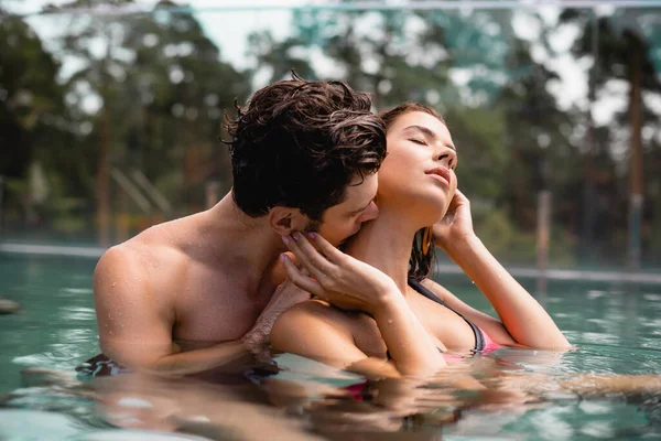 selective focus of muscular man kissing neck of sexy woman in swimming pool