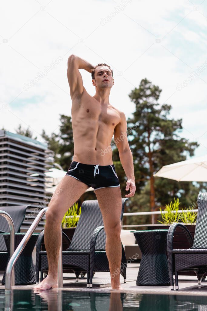 muscular man touching hair while standing near swimming pool 