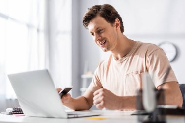 Selective focus of smiling freelancer looking at laptop screen and holding cellphone at home, earning online concept clipart