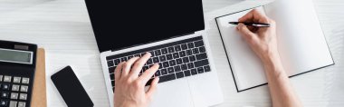 Bir adamın dizüstü bilgisayarda yazarken ve not defterine yazarken panoramik bir kare ve masadaki akıllı telefon, online konsept kazanırken.