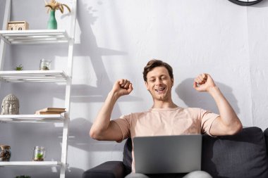 Selective focus of cheerful freelancer showing yeah gesture while looking at laptop on couch at home, earning online concept clipart