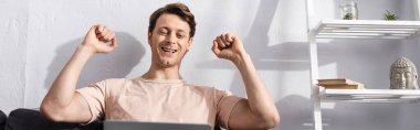 Panoramic crop of happy freelancer showing yes gesture while using laptop at home, earning online concept clipart