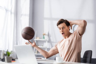 Selective focus of shocked man holding basketball and looking at laptop on table, concept of earning online clipart