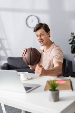 Basketbol tutan neşeli bir adamın seçici odak noktası ve evet jestini masada göstermek, online konsept kazanmak
