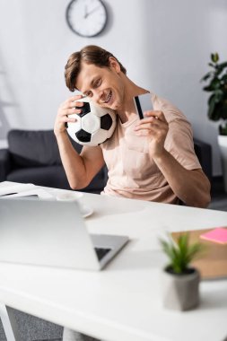 Gülümseyen bir adamın kredi kartını ve futbolu masanın yanında tutarak online konsept kazanmasının seçici odağı.