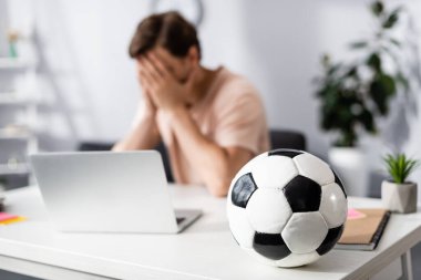 Selective focus of football on table and sad man covering face with hands near laptop at home, earning online concept clipart