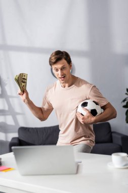 Selective focus of excited man holding cash and football near laptop on table at home, concept of earning online clipart