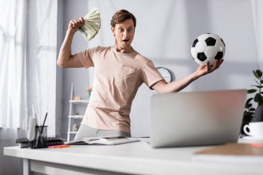 Selective focus of shocked man with cash and football looking at laptop in living room, concept of earning online clipart
