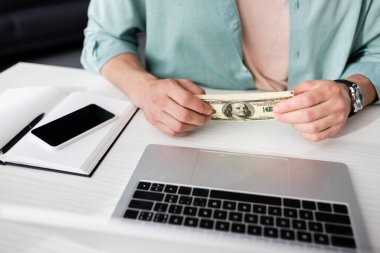 Bilgisayarın yanında dolar tutan adamın görüntüsü ve masada boş ekran olan akıllı telefon, online kazanç kavramı.