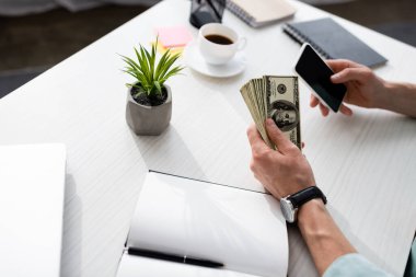 Dolarları elinde tutan, masanın yanında akıllı telefon kullanan ve internet konsepti kazanan bir adam görüntüsü.