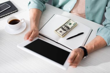 Kahvenin yanında dijital tablet tutan serbest çalışanın görüntüsü ve masadaki defter üzerinde dolar banknotları, online kazanç kavramı.