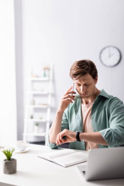 Akıllı telefondan konuşan serbest çalışanın seçici odak noktası ve evdeki kol saati zamanını kontrol etmek, zaman yönetimi kavramı 
