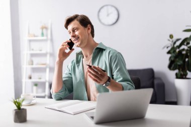 Oturma odasında bilgisayarın ve defterin yanında akıllı telefondan konuşan gülümseyen tele-çalışanın seçici odak noktası, online konsept kazanmak.