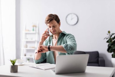 Selective focus of man checking time while talking on smartphone near laptop and notebook on table, concept of time management  clipart